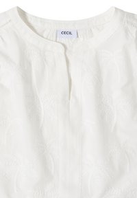 Blouse blanche avec un motif discret de palmier brodé, col rond et petite fente à l'avant, étiquette "Cecil" à l'intérieur du col.