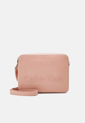 Prostokątna, jasnoróżowa skórzana torba na ramię z wytłoczonym logo Calvin Klein i regulowanym paskiem na jednolitym tle.