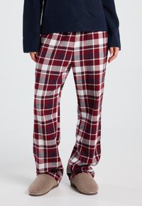 Persoon in rode, witte en blauwe geruite pyjamabroek met beige fuzzy pantoffels en een donkerblauw longsleeve shirt, staande op een witte vloer.
