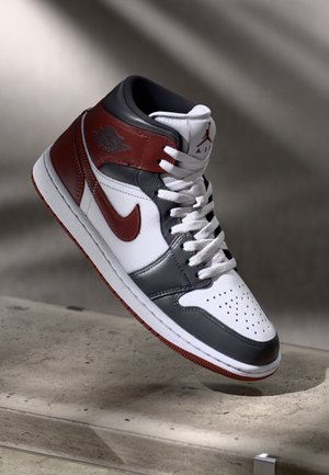 En hvid, grå og mørkerød high-top Air Jordan sneaker med hvide snørebånd, der hviler på en betonoverflade.