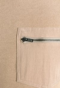 Tessuto beige con superficie strutturata presenta una piccola tasca a toppa, chiusa con una cerniera metallica nera e dettagli di cuciture argentate.