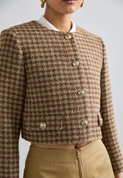 Chaqueta cropped marrón de pata de gallo con botones dorados, tela texturizada y un collar blanco. Combinada con jeans kaki de cintura alta.