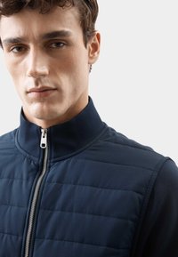 Mørkeblå ærmeløs quilted vest med høj krave og sølv lynlåsdetalje, der viser en glat tekstur og struktureret design.