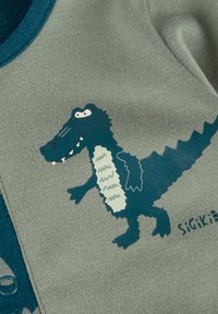 Green cotton garment with a blue crocodile graphic. The crocodile features white accents and a playful expression, set against a textured fabric.
