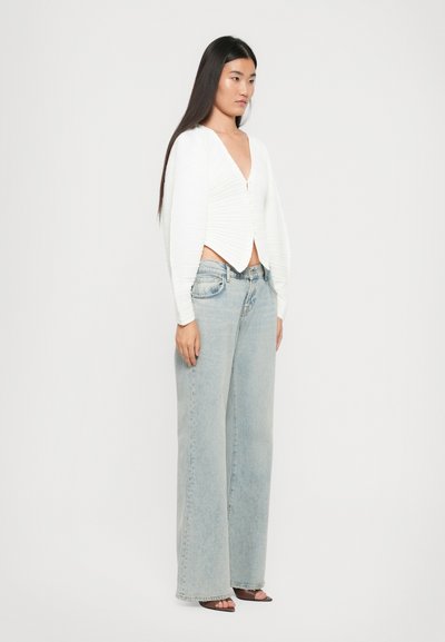Vrouw staat met een witte gerimpelde lange mouwtop, lichtblauwe wijde jeans en bruine open teen hakken voor een effen achtergrond.
