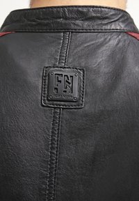 Sort læderjakke med glat tekstur, med en central søm og en fremtrædende firkantet logopatch. Røde accenter på skuldrene.