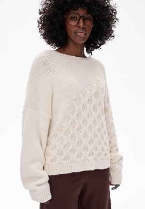 Vrouw met krullend haar die grote bril draagt en een crèmekleurige kabelgebreide trui met gemusterde en effen breisel delen, in combinatie met een bruine broek.