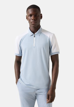Jonge man die een lichtblauw-wit poloshirt met korte mouwen en een ritskraag draagt, staat met één hand in de zak tegen een effen achtergrond.