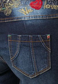 Tummansiniset farkut, joissa on takatasku ja kultaisella brodeeratuksi tekstiksi "We" sekä punainen sydän vyötäröllä.