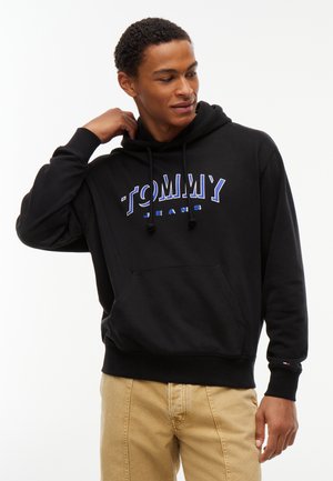 Νέος άνδρας με μαύρη μπλούζα με κουκούλα Tommy Jeans και μπεζ παντελόνια, που ρυθμίζει το γιακά της μπλούζας με το δεξί του χέρι, κοιτάζοντας στο πλάι.