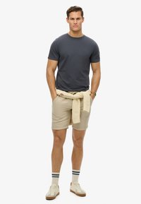 Camiseta gris de cuello redondo combinada con pantalones cortos beige. Un suéter de punto color crema está atado a la cintura. Zapatillas blancas con detalles en marrón.