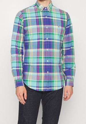 Homme portant une chemise à manches longues à carreaux bleus, verts, blancs et violets et un pantalon noir.