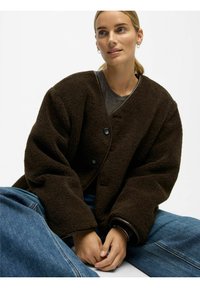 Braune, übergroße Fleecejacke mit strukturierter Oberfläche, ausgestattet mit einem tiefen V-Ausschnitt, großen Knöpfen und kontrastierendem Besatz. Wird über einem grauen Oberteil getragen.