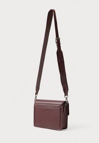 Brązowa skórzana torba na ramię z regulowanym paskiem i zapięciem na klapkę, zawieszona na białym tle.