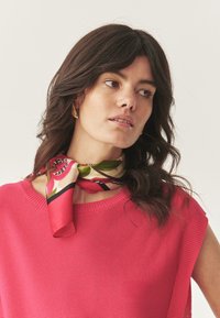 Top rosa lavorato a maglia con maniche corte, abbinato a una sciarpa di seta ornamentale con disegni floreali nei toni del rosa e del verde.