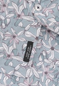 Bloes met bloemen op een lichtblauwe achtergrond, versierd met witte, roze en turquoise bloemen, en een zwart label met de tekst "IEDERE STAP EEN NIEUWE REIS."