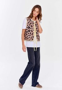 Žena stojící, oblečená v růžovém a černém leopardím vzoru, má na sobě vestu přes bílý tričko, tmavé džíny a hnědé tenisky, na čistém bílém pozadí.