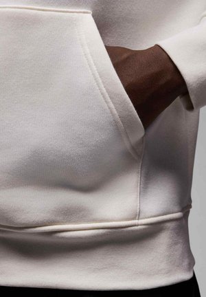 Main à la peau foncée partiellement dans la poche avant d'un sweat-shirt couleur crème, montrant la texture du tissu et les détails de la couture.