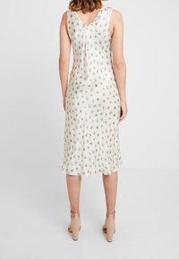 Robe sans manches crème avec un motif floral vert, design dos en V, qui tombe jusqu'au mi-mollet, fabriquée en tissu lisse, avec un ourlet évasé.