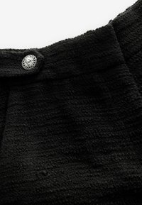 Tissu noir texturé avec un motif subtil, comportant un bouton décoratif en argent à la taille. Gros plan sur le matériau et les détails.