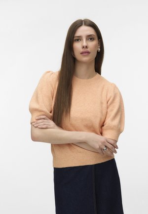 Kobieta z długimi, prostymi brązowymi włosami, ubrana w brzoskwiniowy sweter z krótkim rękawem i ciemną spódnicę, stoi z założonymi rękami na tle o jednolitym kolorze.