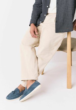 Person, die auf einem hölzernen Hocker sitzt, trägt cremefarbene Hose, schwarzes kariertes Hemd und blaue Wildleder-Slipper mit Quasten.