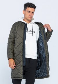Quiltierter olivgrüner langer Mantel mit Reißverschluss, der ein dunkles Innenfutter aufweist und offen über einem weißen Hoodie mit bedrucktem Text getragen wird.