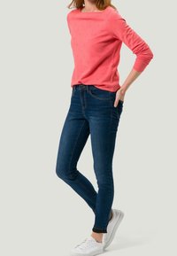 Suéter de punto color coral con patrón texturizado, combinado con jeans ajustados de un azul oscuro y zapatillas blancas. Cuello redondo simple y silueta ajustada.