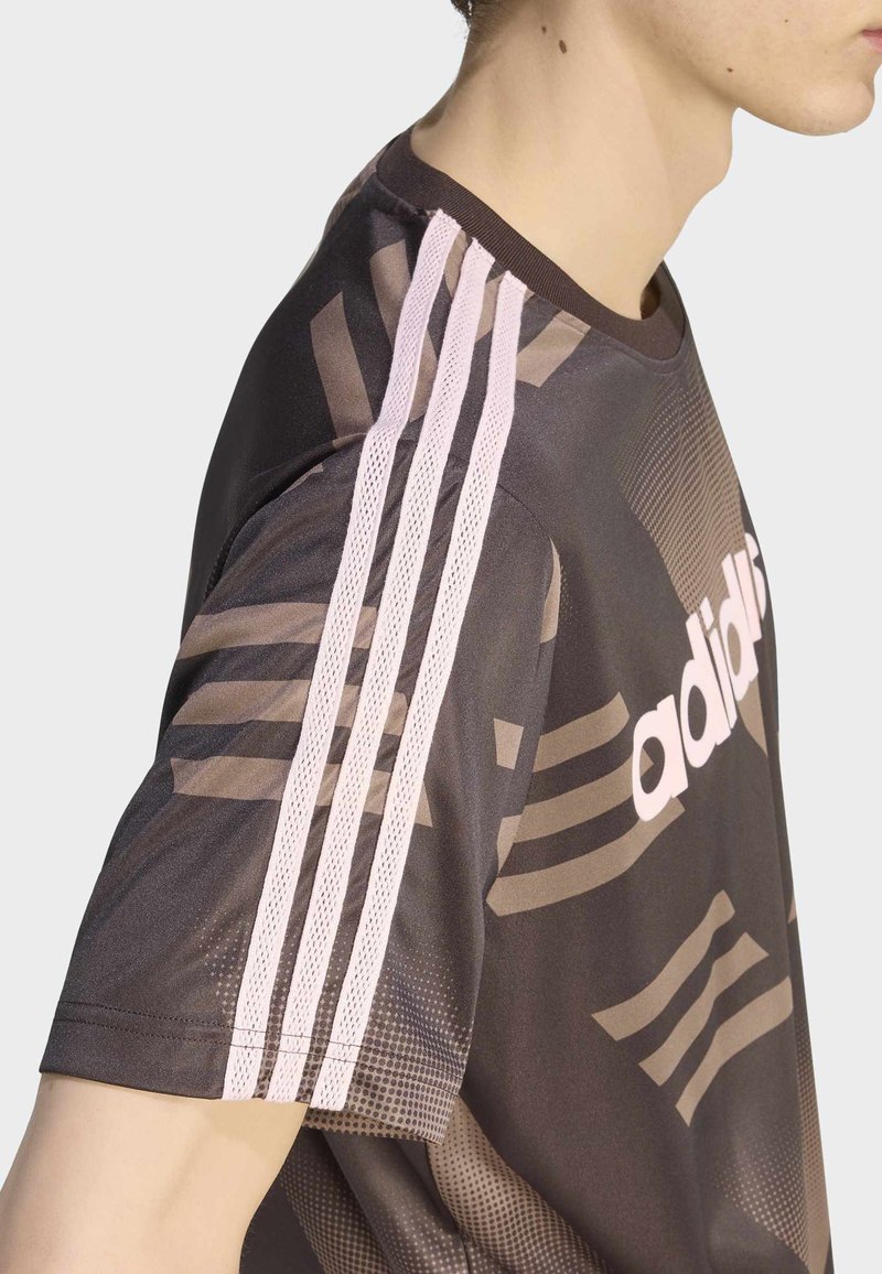 Uomo che indossa una maglia Adidas marrone scuro con tre strisce rosa chiaro sulla spalla e logo parziale Adidas sul petto.