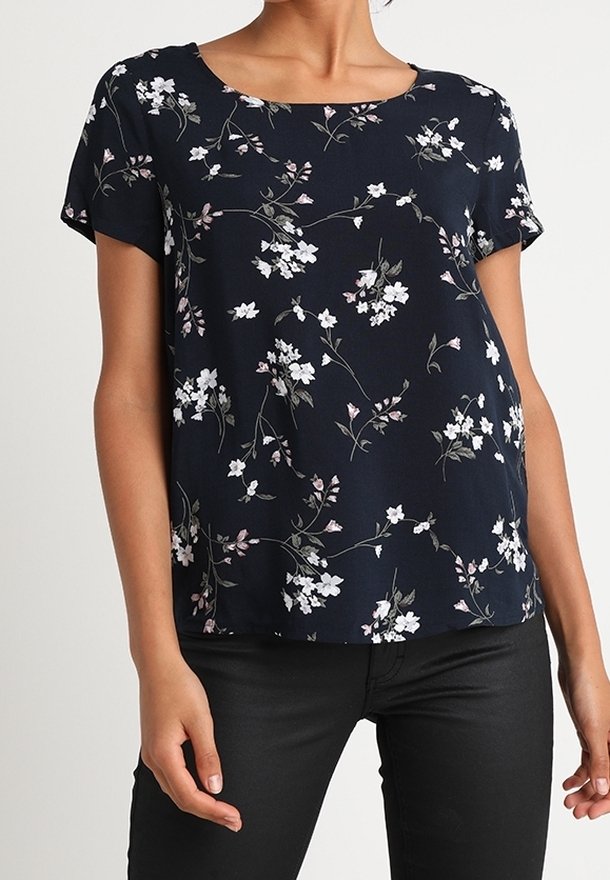 Femme portant un chemisier bleu marine à manches courtes avec un imprimé floral blanc et rose, associé à un pantalon noir.