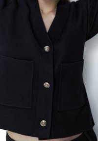 Navy knitted cardigan featuring a V-neck, two front pockets, and decorative gold buttons with intricate designs. Soft texture and fitted style.