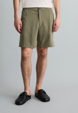 Man draagt olijfgroene casual shorts, wit overhemd, zwarte instapschoenen, staand tegen een effen grijze achtergrond.