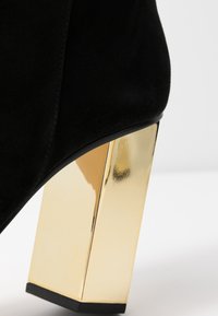 Botín de ante negro con un tacón cuadrado grueso y brillante en color dorado, que presenta una textura suave y un diseño de punta redondeada.