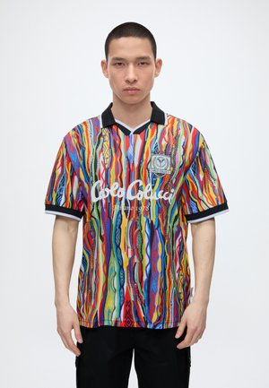 Tânăr cu păr scurt purtând un tricou polo cu dungi multicolore vibrante, cu guler și mâneci negre, stând pe un fundal alb simplu.