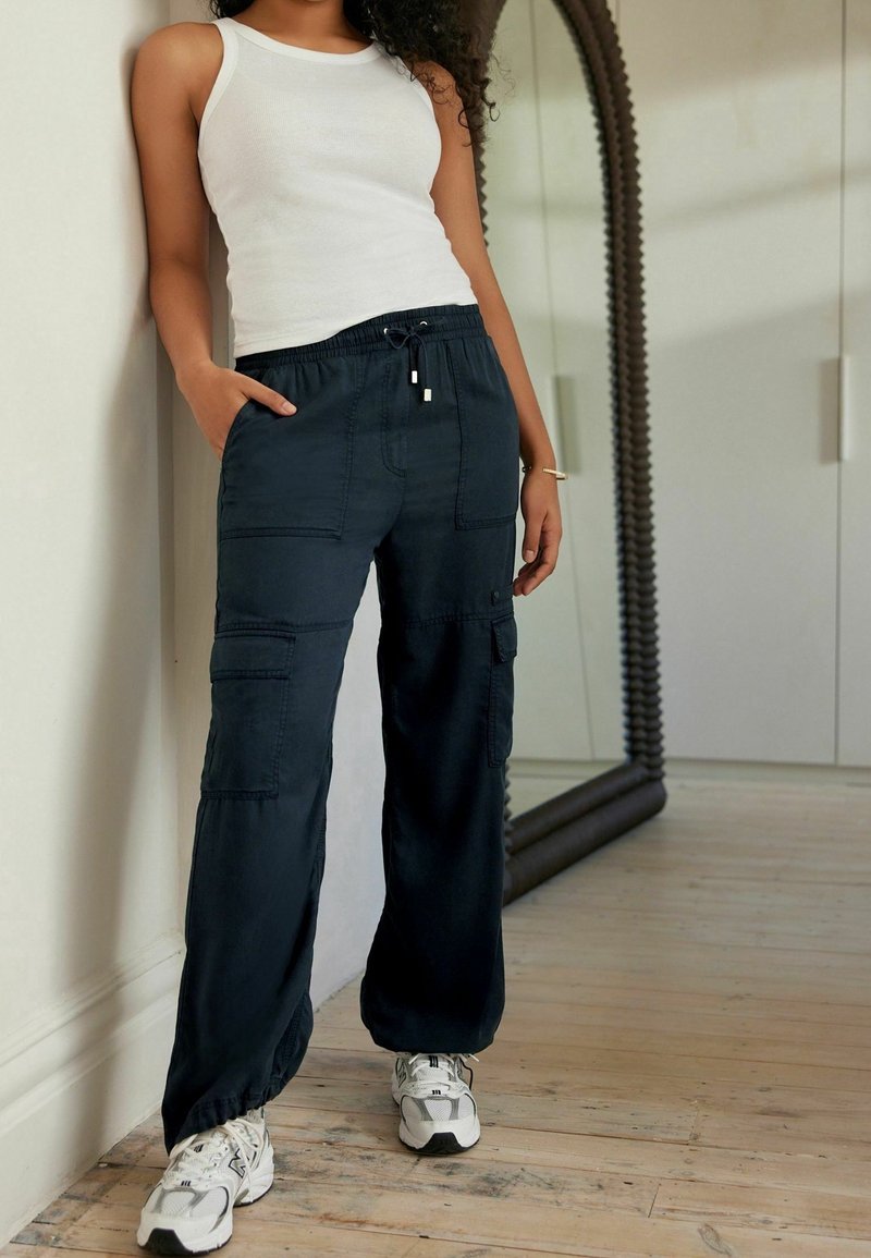 Femme portant un débardeur blanc, un pantalon cargo noir et des baskets blanches, debout à l'intérieur près d'un grand miroir et d'un sol en bois.