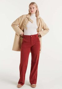 Gabardina beige con acentos florales, cuello de tortuga blanco y pantalones de pana rojos. La modelo está de pie con las manos en las caderas, vistiendo zapatos de color beige claro.