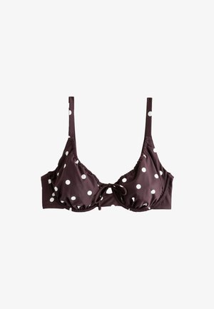 Femme portant un bikini marron à pois blancs, debout au soleil avec les mains dans le dos.