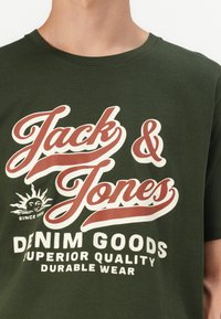 Alyvuogių žalios spalvos medvilnės marškinėliai su drąsiais baltais ir raudonmedžio spalvos užrašais. Dizainą papildo saulės grafika ir tekstas "DENIM GOODS, SUPERIOR QUALITY."