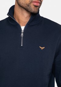 Marineblauwe rits sweatshirt met hoge kraag, gemaakt van zacht materiaal. Voorzien van een zilveren rits en een geborduurd gouden logo op de borst.
