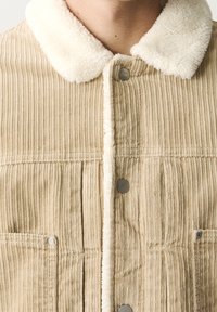 Beige kordflöjlsjacka med en vit faux shearling-krage, som har två fickor fram och metallknappar längs placket. Texturerat material.