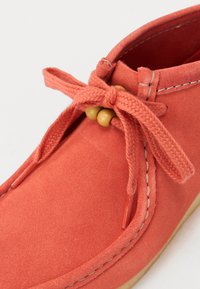 Zapato de ante coral con punta redonda, cordones trenzados y detalle de cuentas de madera, con acentos de costura a lo largo del borde de la parte superior.