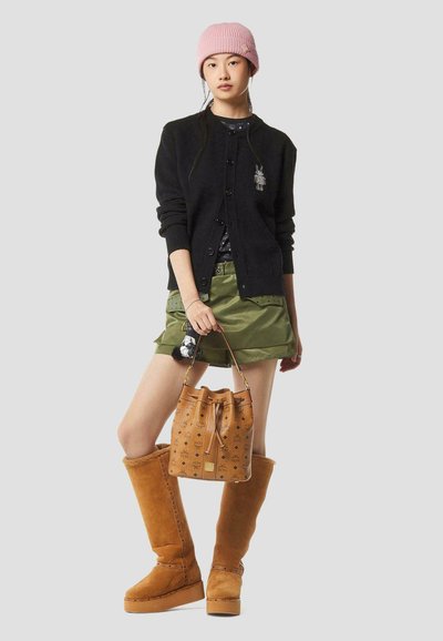 Jonge vrouw met een roze muts, een zwart vest, groene korte broek, beige kniehoge laarzen, en een beige gestreepte emmerentas vasthoudend.