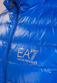 Niebieska puchowa kurtka z błyszczącą, pikowaną teksturą, z zamkiem błyskawicznym i srebrnym logo EA7 Emporio Armani na piersi.