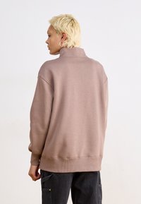 Felpa marrone over size con colletto alto, polsini e orlo a costine. Tessuto liscio, realizzata in stoffa morbida, progettata per il comfort.