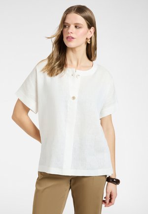 Vrouw die een wit blouse met korte mouwen en knopen draagt, met beige broek, gouden hoepeloorbellen en een donkere armband, kijkt naar links.