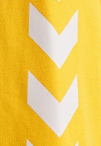 Żółta tkanina z wzorem trzech dużych białych, skierowanych w dół szewronów ułożonych pionowo.