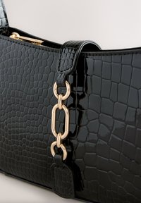 Mini-sac à main en cuir verni noir avec un motif crocodile texturé, agrémenté d'une chaîne dorée et d'une fermeture éclair sur le dessus.