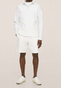 Felpa bianca con zip e maniche lunghe abbinata a pantaloni shorts bianchi. Texture liscia, stile casual, indossata con sneaker bianche.