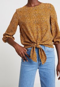 Senfbluse mit weißen und schwarzen Punkten, Dreiviertelärmel, Knoten-Frontdetail, kombiniert mit hellblauen Jeans.