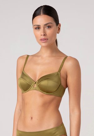 Donna con capelli scuri legati indossa reggiseno di raso verde oliva e biancheria intima coordinata su sfondo chiaro uniforme.